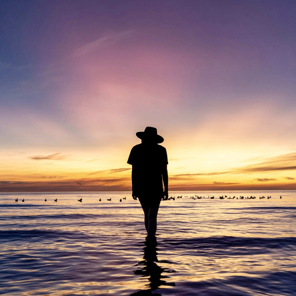 Woman in hat silhouetted against sunset, walking into calm ocean water
