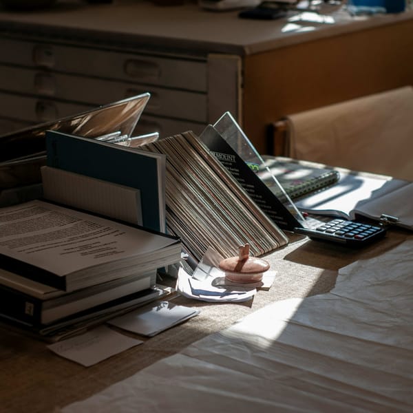 Cluttered desk with stacked documents, folders, and a calculator in dim afternoon light, chair empty